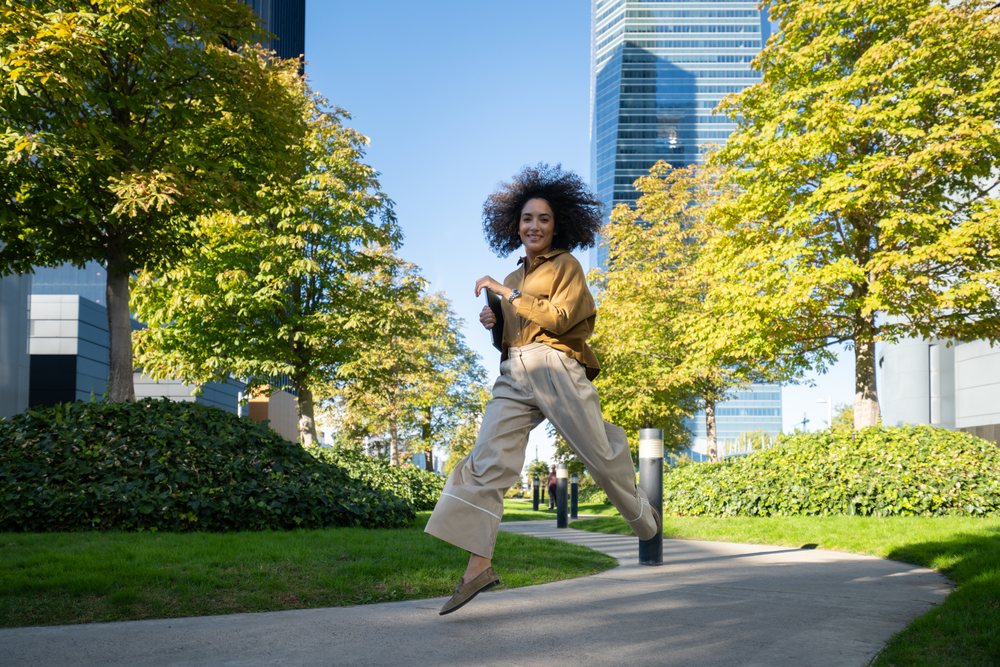 Woman joyfully jumps on a sidewalk in an urban park with trees and skyscrapers on a sunny day, Madrid, Spain.