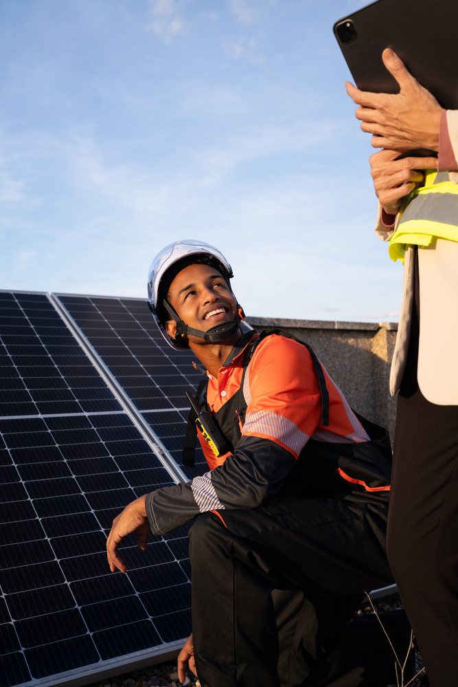 Worker wearing a helmet inspects solar panels on a rooftop in Madrid while another person holds a tablet device nearby under a blue sky, Spain.