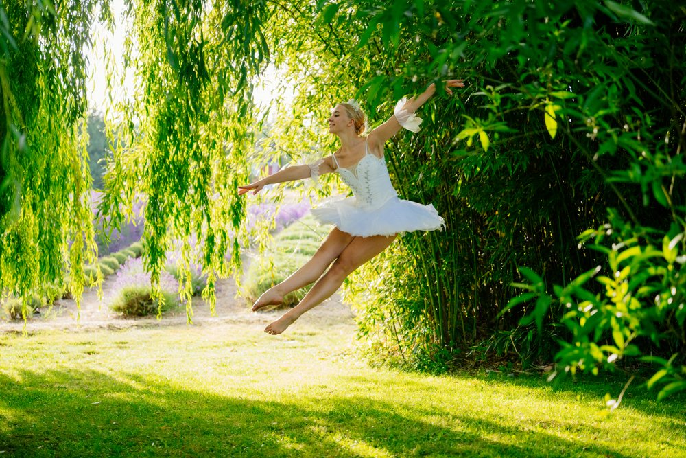 A ballerina in a white tutu leaps gracefully outdoors surrounded by lush greenery and willow trees, WA, USA.
