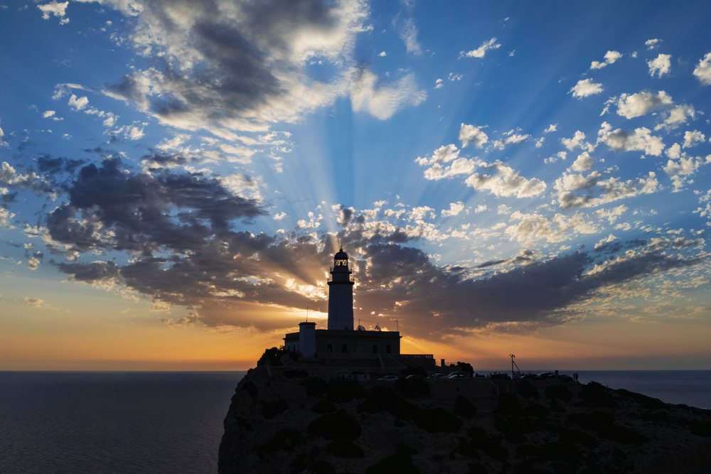 Silhouetted Far de Formentor Lighthouse with sun rays breaking through clouds at sunrise on Mallorca's Cap de Formentor coast, Landscape, Lifestyle, Polença, Spain.