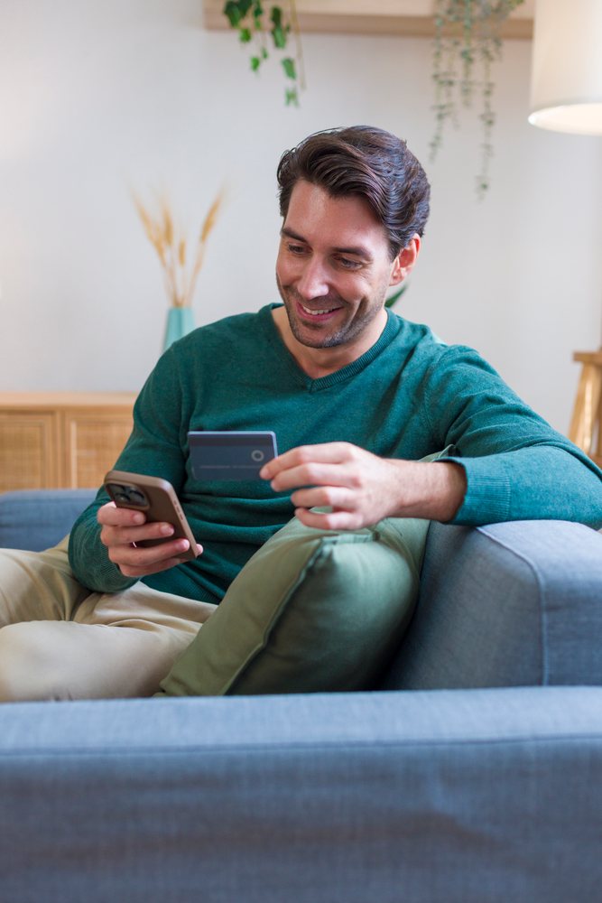 Man sits on a sofa holding a credit card and smartphone in a modern home interior with a green pillow nearby, Technology, young adult.