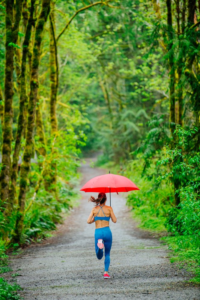 Woman running on a wet gravel forest path holding a red umbrella during rainy weather, Calisthenics, USA.