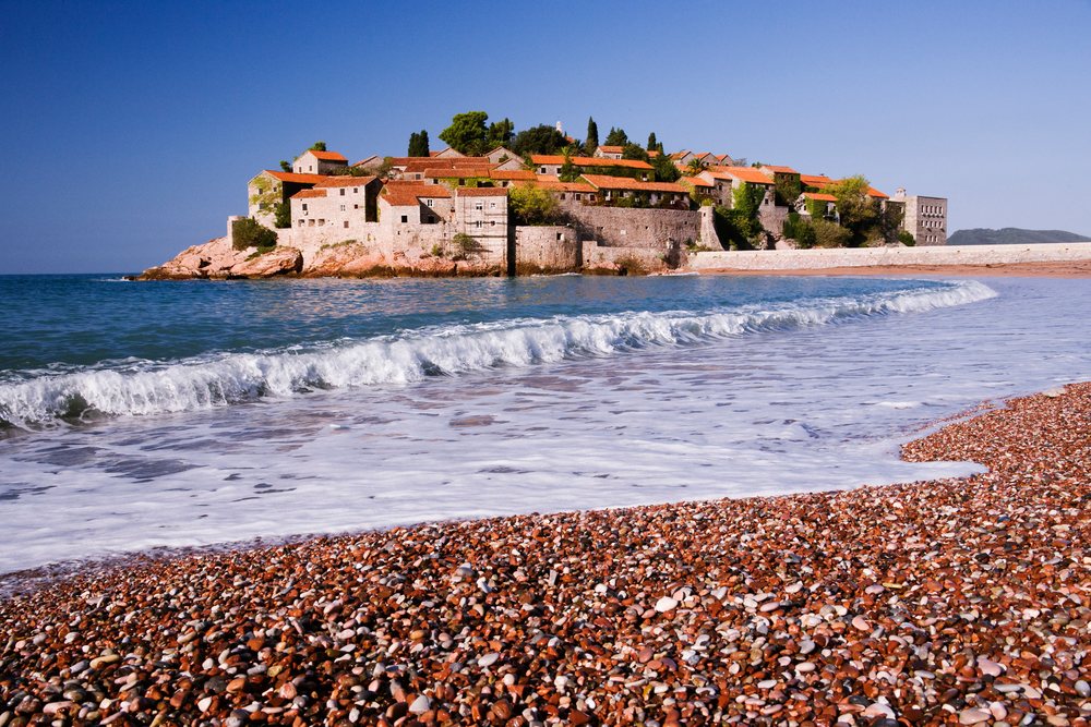 Pebble beach with waves and the historic island village of Sveti Stefan under a clear blue sky.