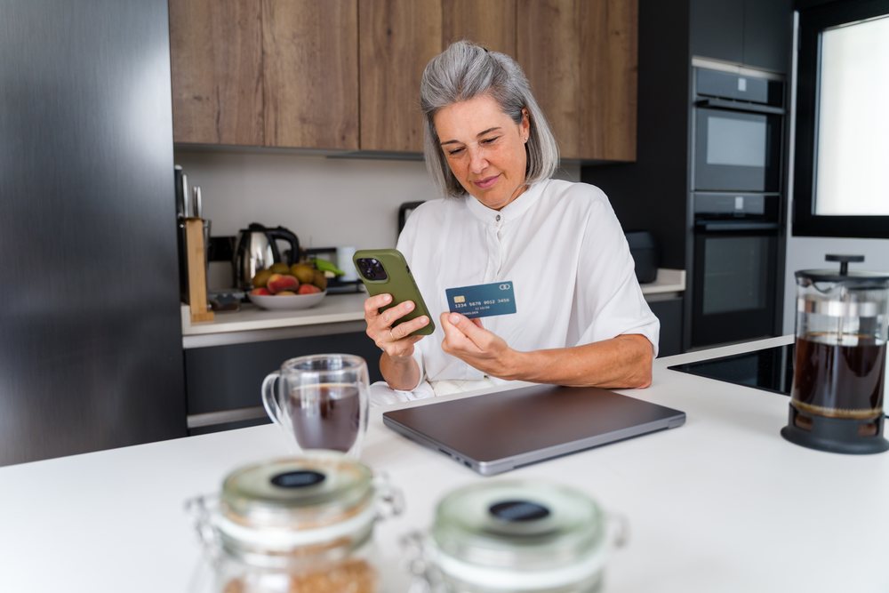 Woman sitting in a modern kitchen in Spain using a smartphone and holding a payment card for online shopping.