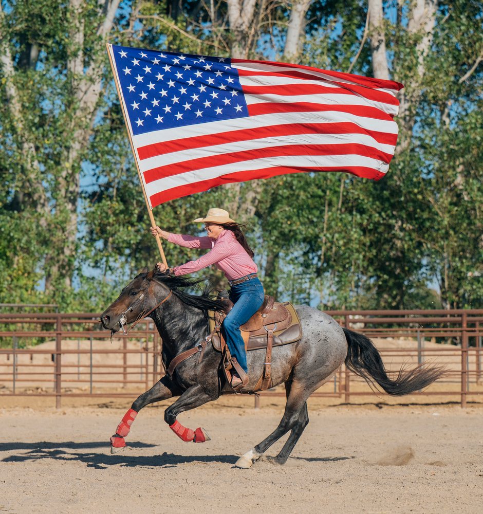 Woman wearing cowboy hat rides horse holding large American flag in a fenced outdoor western riding arena, Southwest USA.