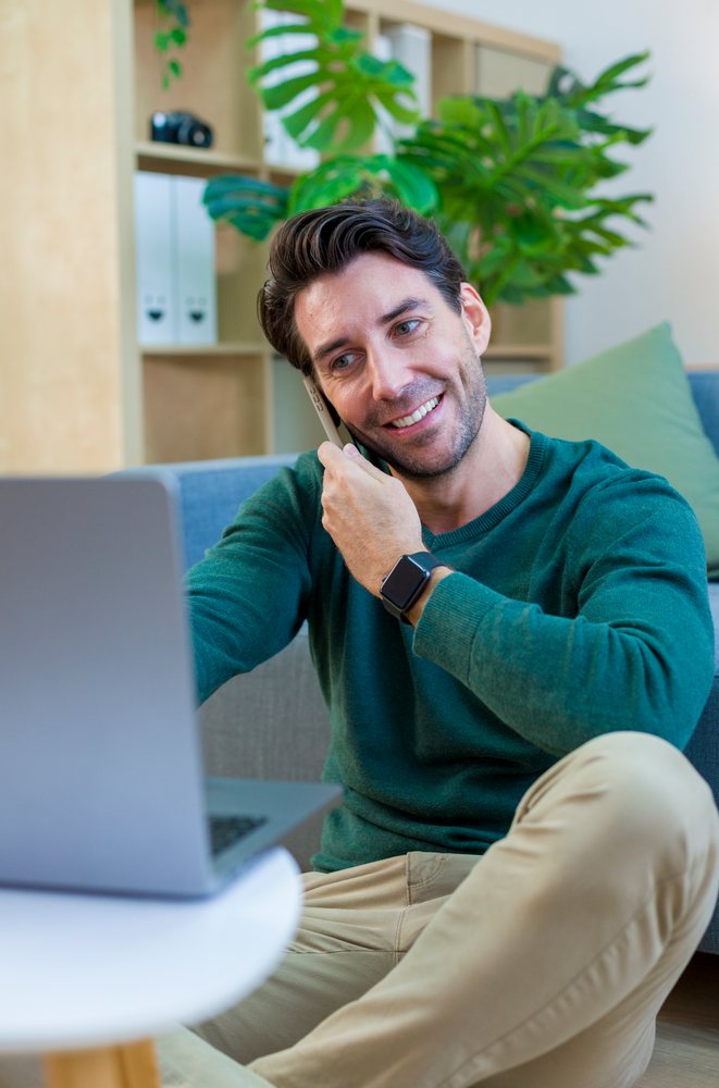 Man sitting on the floor using a laptop and talking on the phone in a modern home office, technology, young adult.