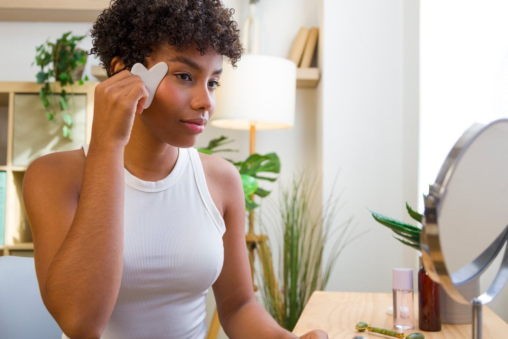 Young woman performs facial massage using a tool in front of a mirror during her home skincare routine.