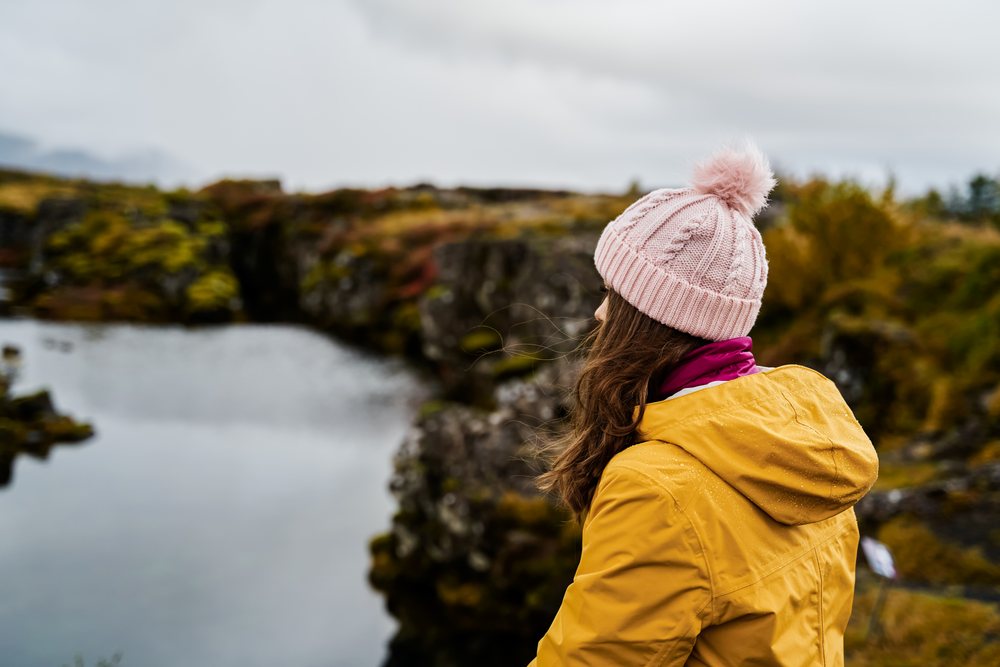 A woman wearing a yellow outerwear and pink knit headwear looks over a moss-covered rocky shore and lake on a cloudy day, Iceland.