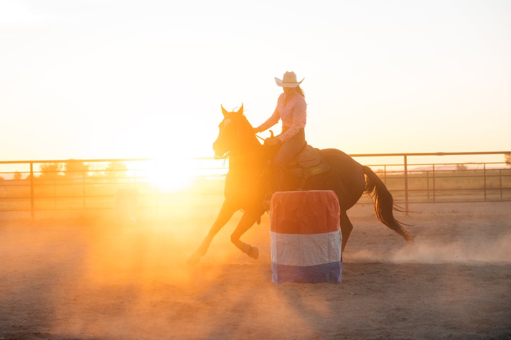 Woman wearing cowboy hat rides horse around barrel in dusty outdoor arena at sunset, Southwest USA.