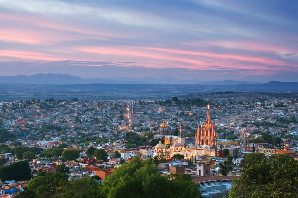 Cityscape of San Miguel de Allende at sunset with illuminated buildings, a church cross, and mountains in the background, Mexico.