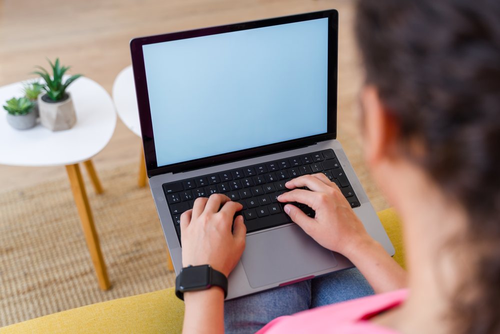 A person types on a laptop keyboard while sitting indoors at home with a small table holding potted plants nearby, Spain.