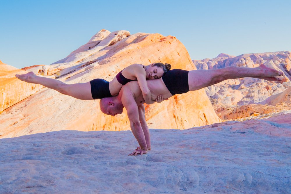 Man supports a woman performing acrobatic yoga on rocky terrain outdoors under a clear blue sky, USA.