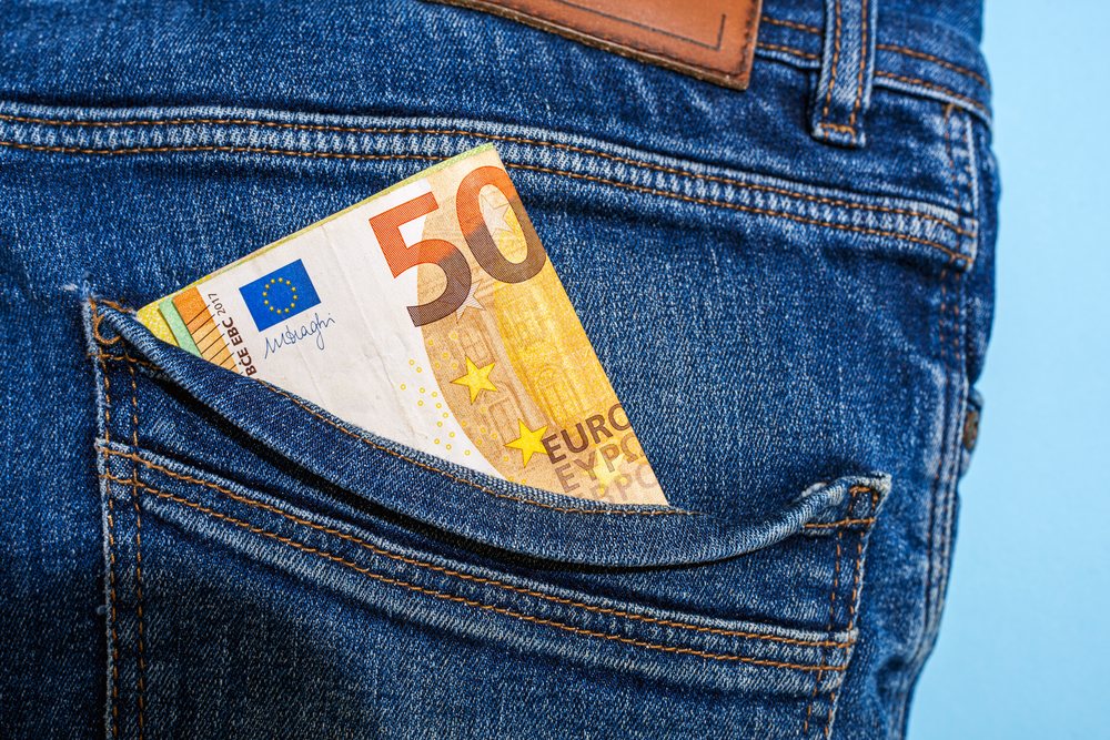 50 Euro banknote partially visible in the back pocket of blue denim jeans against a plain blue background in studio lighting, Studio Shot.
