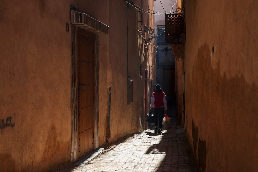 Man walking away carrying grocery bags down a narrow, sunlit alleyway with textured earthen walls and cobblestone path, Lifestyle, Landscape, Culture, Marrakech, Marrakesh-Safi Region, Morocco.