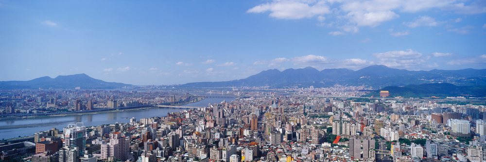 Urban cityscape featuring a river and bridges beneath a blue sky with scattered clouds and distant mountains.