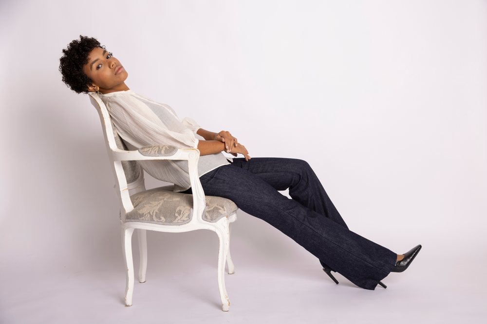 Curly-haired woman wearing white blouse and dark jeans sits relaxed, leaning back on an ornate white chair in a studio with white background, fashion, young culture.