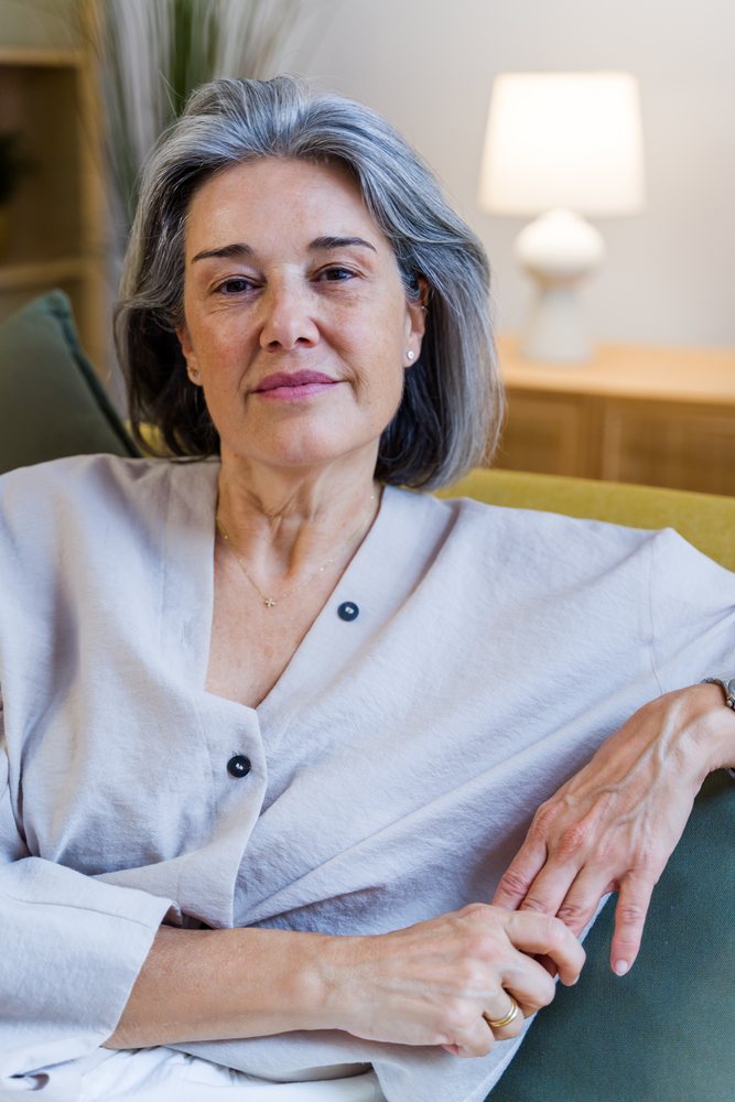 Mature woman with gray hair sitting relaxed on a sofa in a calm indoor setting with wooden cabinet and lamp, Spain.