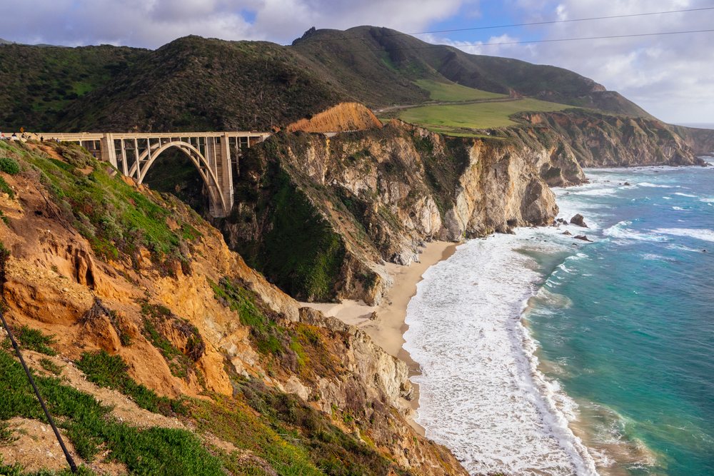 The Bixby Bridge, a concrete arch, spans coastal cliffs with green vegetation along the California coastline under a partly cloudy sky, USA.