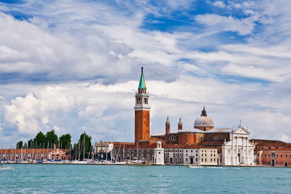 San Giorgio Maggiore church with bell tower and dome on an island, with boats moored on waterfront under a partly cloudy sky, Venice, Italy.