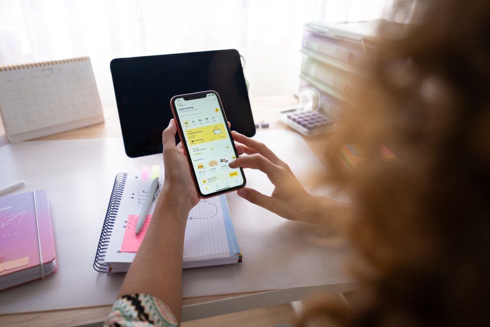 Woman using smartphone app at desk with tablet and notebooks in a home office, Spain.