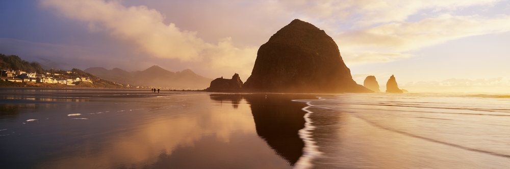 Haystack Rock at sunset with ocean waves, wet reflective sand, and a coastal town, Oregon, USA.