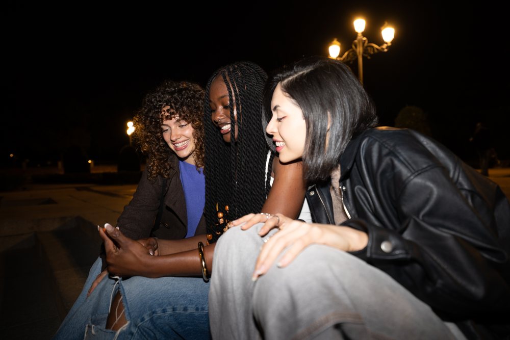 Three women outdoors at night in Madrid smiling and looking at a phone together under streetlights, Spain.