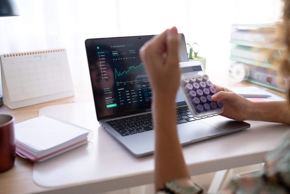 Woman using a calculator and laptop displaying stock market data while working at a home office desk, Spain.