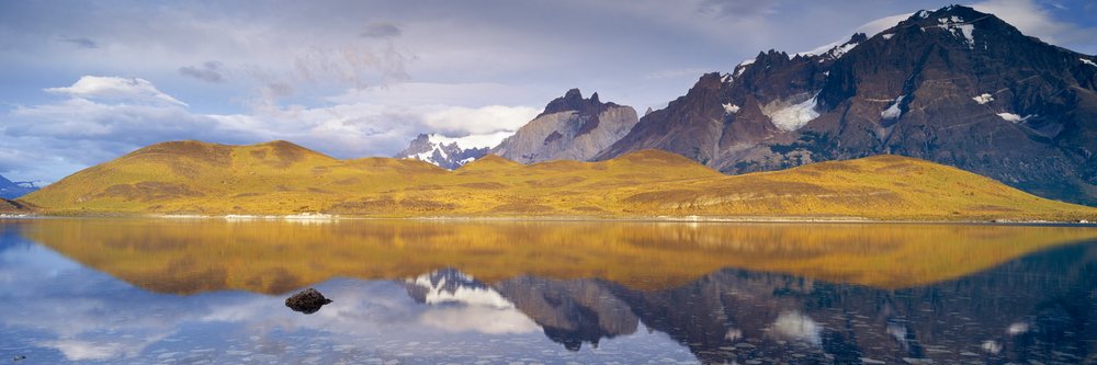 Los Cuernos mountains with snow caps reflect in a calm lake with rocks under a cloudy sky, Torres del Paine National Park, Chile.