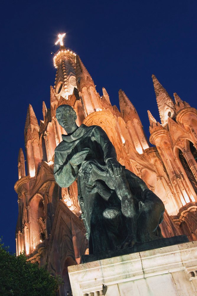 Robed figure statue on a stone pedestal stands before the illuminated gothic La Parroquia church at night, Guanajuato, Mexico.