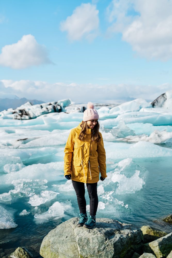 A woman wearing winter clothing stands on a rock by icy glacial water at a glacier lagoon, Iceland.