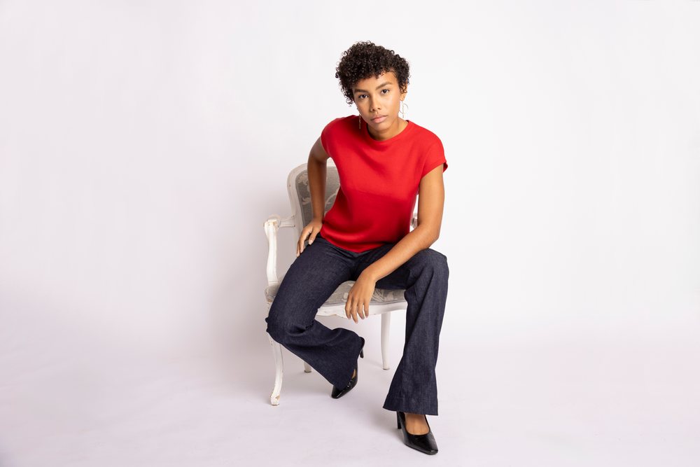 Woman seated on a vintage style chair wearing a red shirt, black pants, black heels, and hoop earrings against a white studio background, fashion, clothes, young culture.
