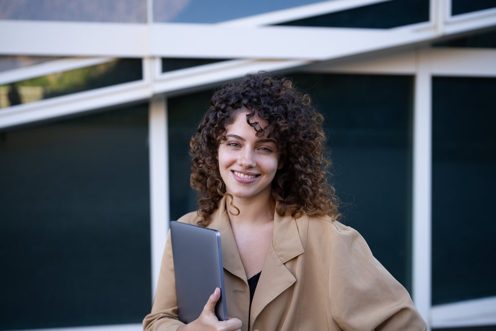Woman with curly hair smiling and holding a laptop outdoors near a modern building window, Spain.