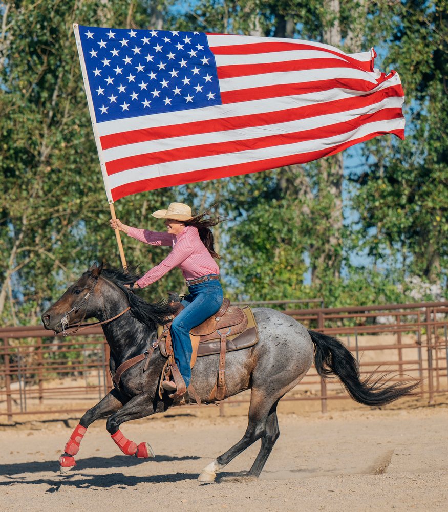Woman wearing cowboy hat and pink shirt rides horse with red leg wraps in dusty rodeo arena holding large American flag, Southwest USA.