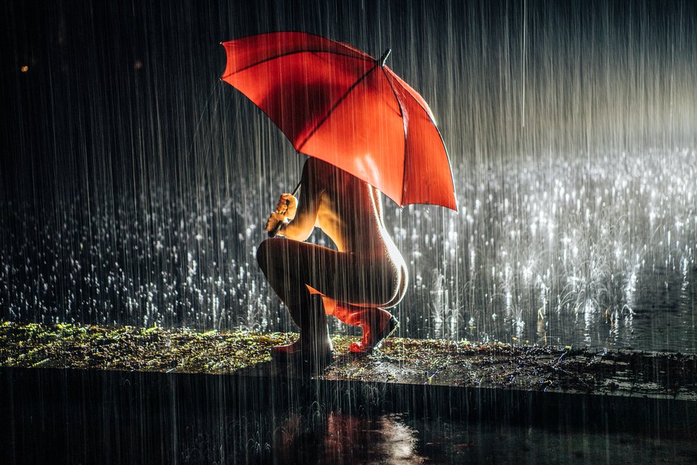 Woman crouching outdoors on wet ground under a red umbrella in heavy rain at night with reflections on the surface, cosplay, nudity, freedom, expression, USA.