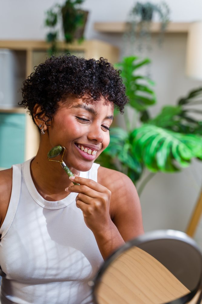 Woman with curly hair smiling while using a jade face roller on her cheek in a bright indoor room with plants and a mirror, Spain.