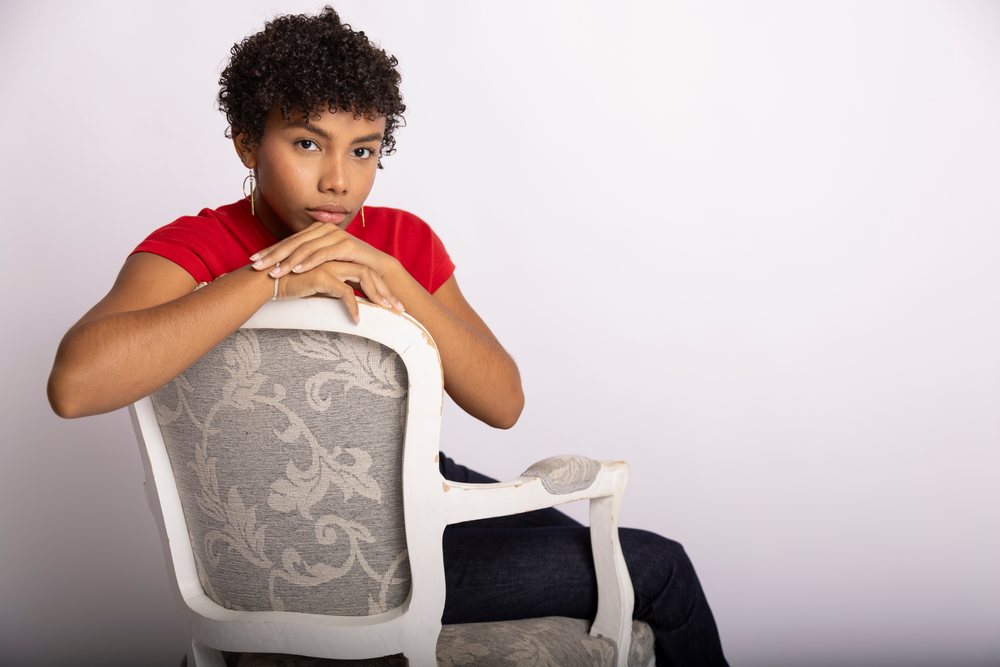 Young woman with curly hair wearing a red shirt and jeans sits on a patterned armchair in a studio, fashion, clothes, young culture.