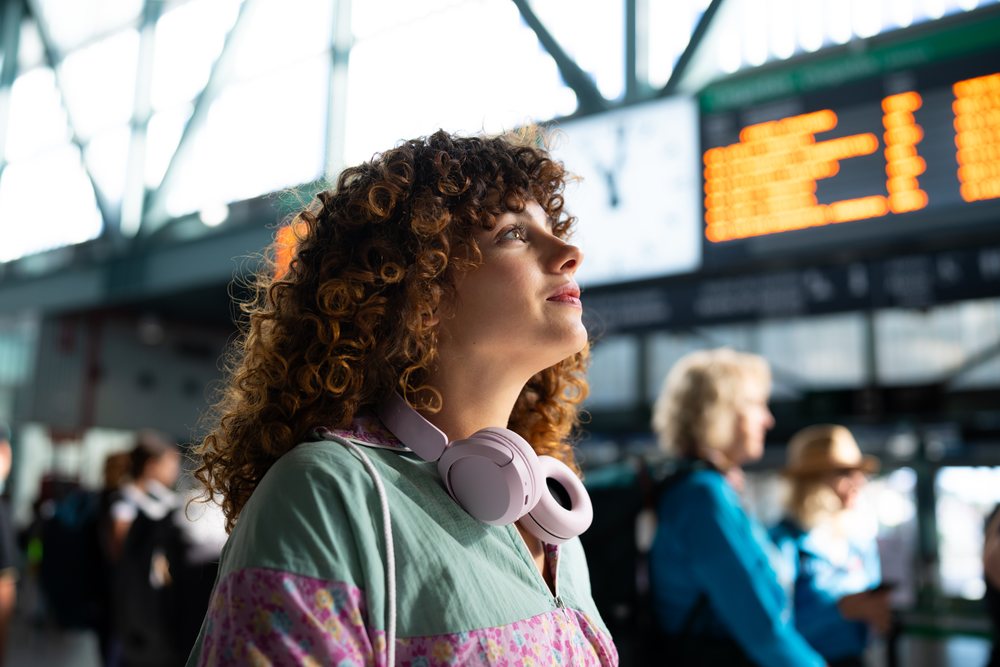 Woman with curly hair wearing pink headphones looks up at the electronic arrival board inside a busy indoor train station, Spain.