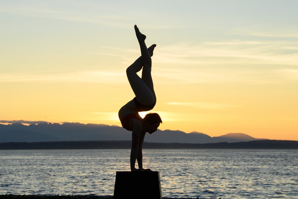 Woman performs a handstand balancing on a box at sunset by the ocean with mountains in the background, USA.