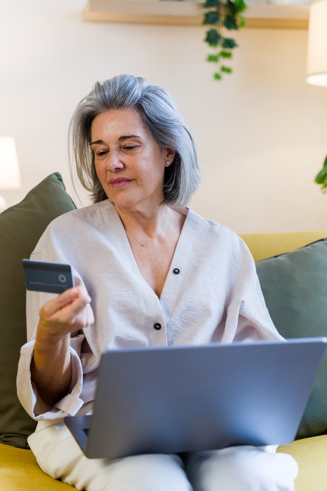 Mature woman with gray hair seated on a yellow sofa holding a credit card and using a laptop indoors at home, Spain.