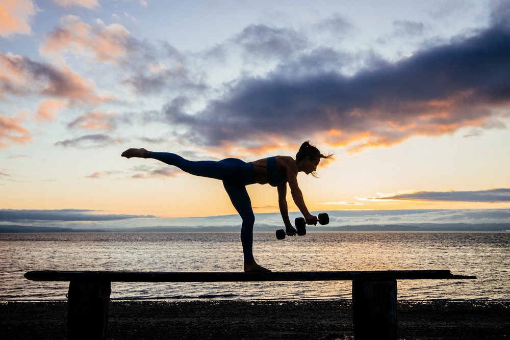 Woman balancing on one leg on a wooden beam holding dumbbells at sunset on a beach, WA, USA.