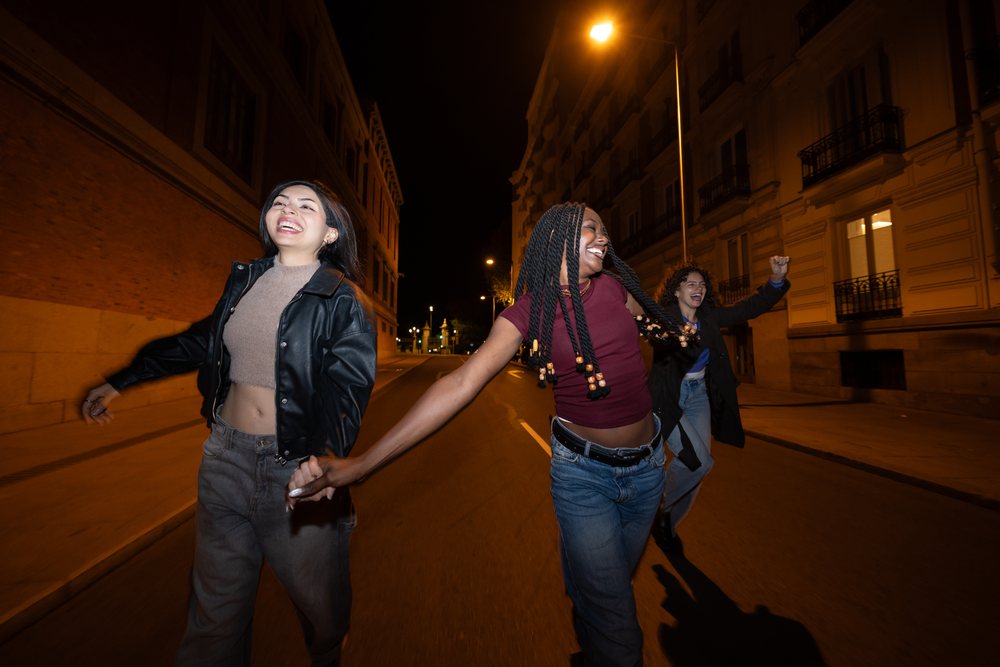 Three women laughing and holding hands while walking on a city street at night, Madrid, Spain.