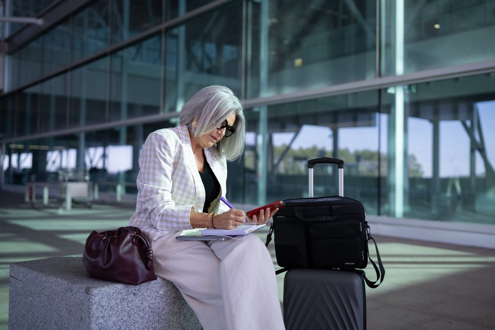 Businesswoman with gray hair writing in a notebook and using a smartphone while sitting outdoors near luggage by a modern building, Spain, Madrid.
