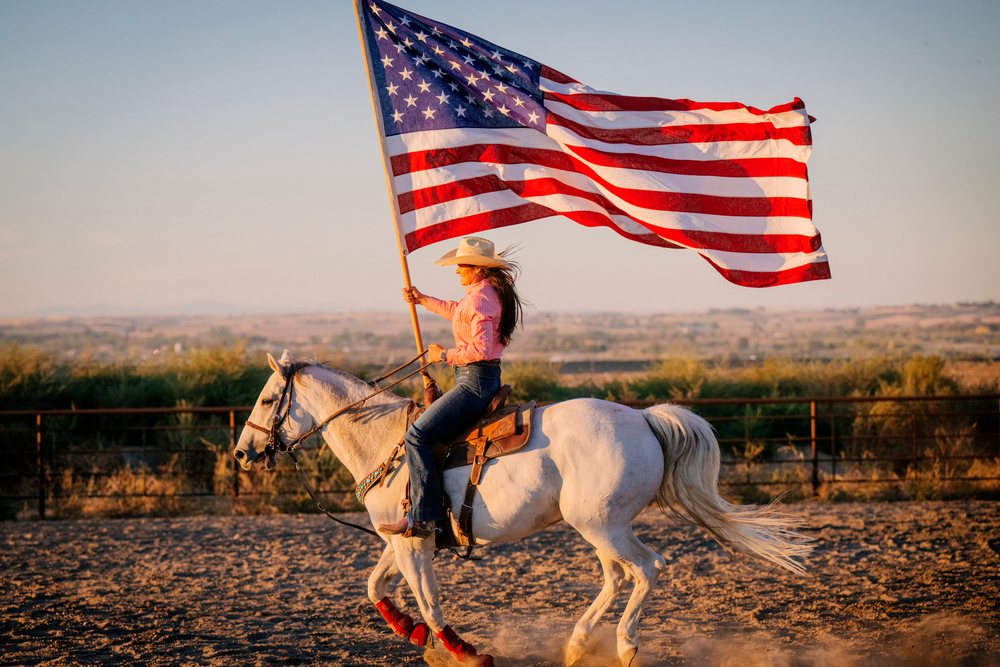 Woman riding a white horse holding a large American flag in a fenced rodeo arena at sunset, Southwest USA.