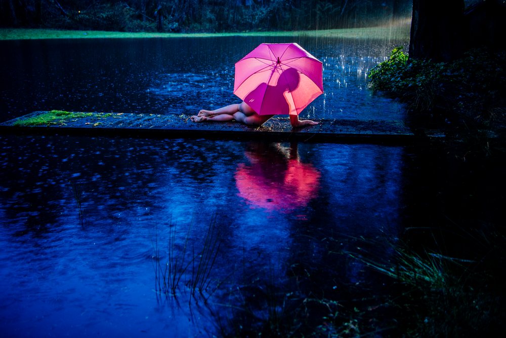 Woman lying on a pathway near water under a pink umbrella in the rain at night with reflections on the wet surface, cosplay, nudity, freedom, expression, USA.