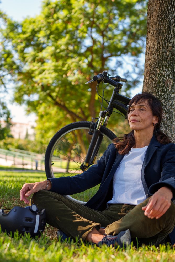 Woman meditating cross-legged against a tree in a grassy park with bicycle and helmet beside her, Málaga.