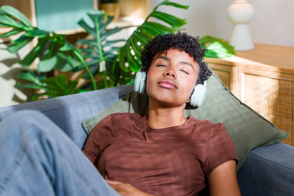 Woman with curly hair wearing headphones relaxes on a sofa indoors beside green plants and a green pillow, sunlight illuminating her face, Spain.