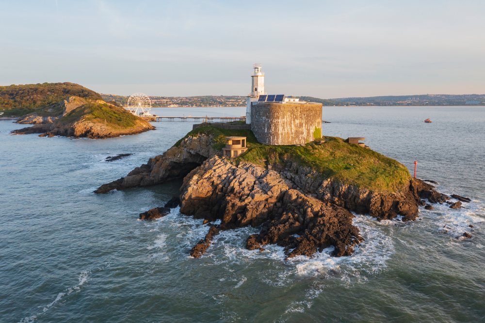 Mumbles lighthouse stands on a rocky island with waves, a pier, and a ferris wheel nearby at Sunrise, Landscape, Swansea, Glamorgan, Wales.