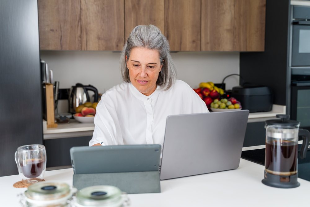 Mature woman multitasking with a tablet and laptop in a modern kitchen with coffee and fruit bowl, Spain.