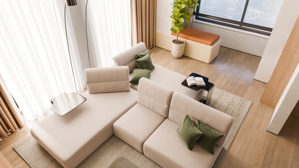 Beige sectional sofa with green pillows beside a black side table holding an open book, near a large window with sunlight and a potted plant.