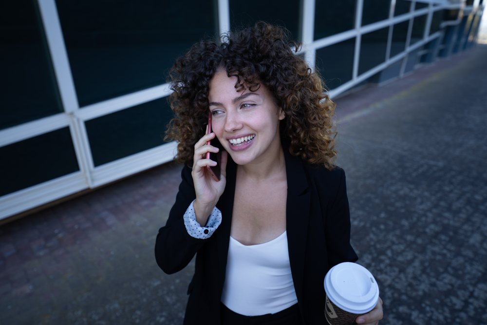 Businesswoman with curly hair talking on phone and holding a coffee cup outdoors on a sidewalk near a modern building, business trip, Spain, Santiago De Compostela.